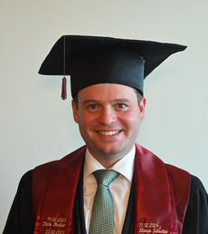 Absolvent in Hochschulrobe mit Doktorhut, rotem Stola und weißem Hemd; Name/Details auf der Stola sichtbar.